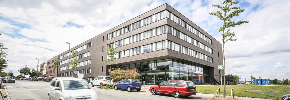 ITMC: Ein großes, braun-rot verklinkertes Gebäude mit Flachdach aus der Froschperspektive fotografiert. Davor eine Straße, auf der ein Auto fährt. Andere Autos stehen am Straßenrand hintereinander geparkt.